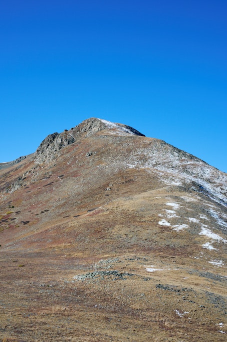 a pointy mountain peak colored with yellow and red plants, dusted with snow