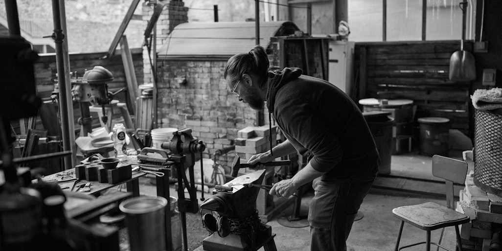 David Bradley, blacksmith hammering a spoon in his work space, sunlight filtering into the outdoor studio