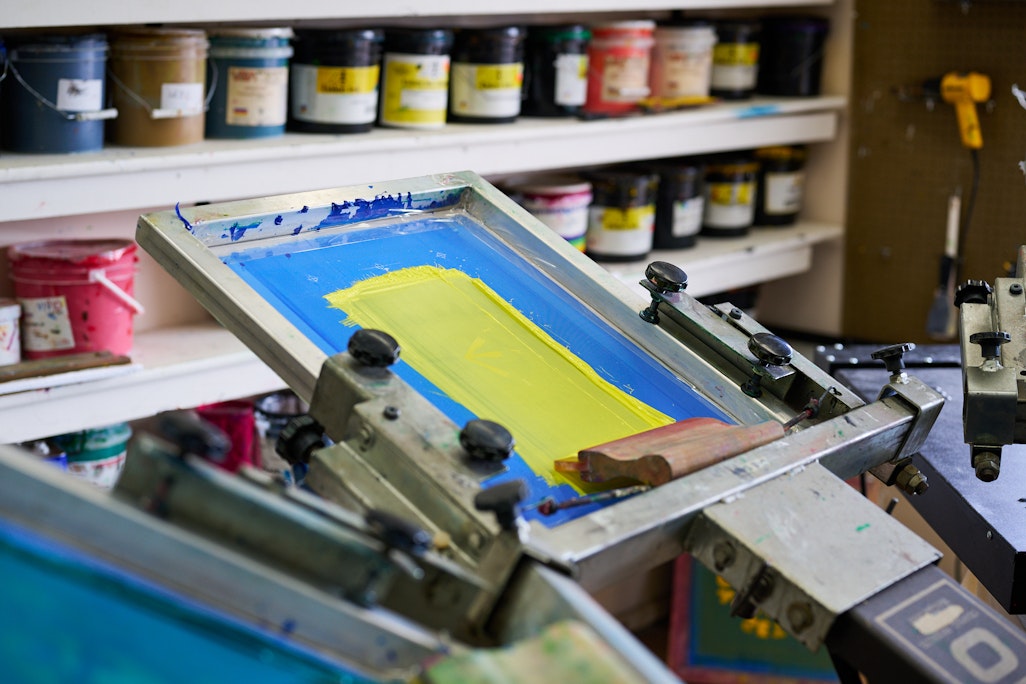 a manual screen printing press, primary colors of blue and yellow, with paint cans in the background of the studio