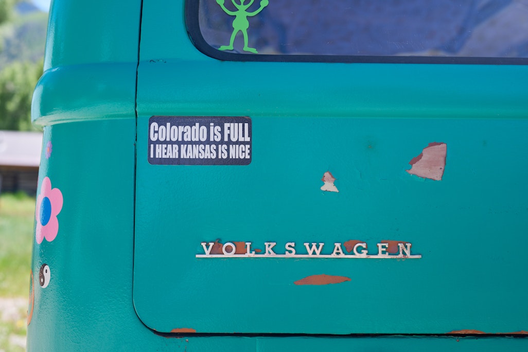 rusty rear view of a green Volkswagen Bus, bumper sticker says “Colorado is FULL I HEAR KANSAS IS NICE”