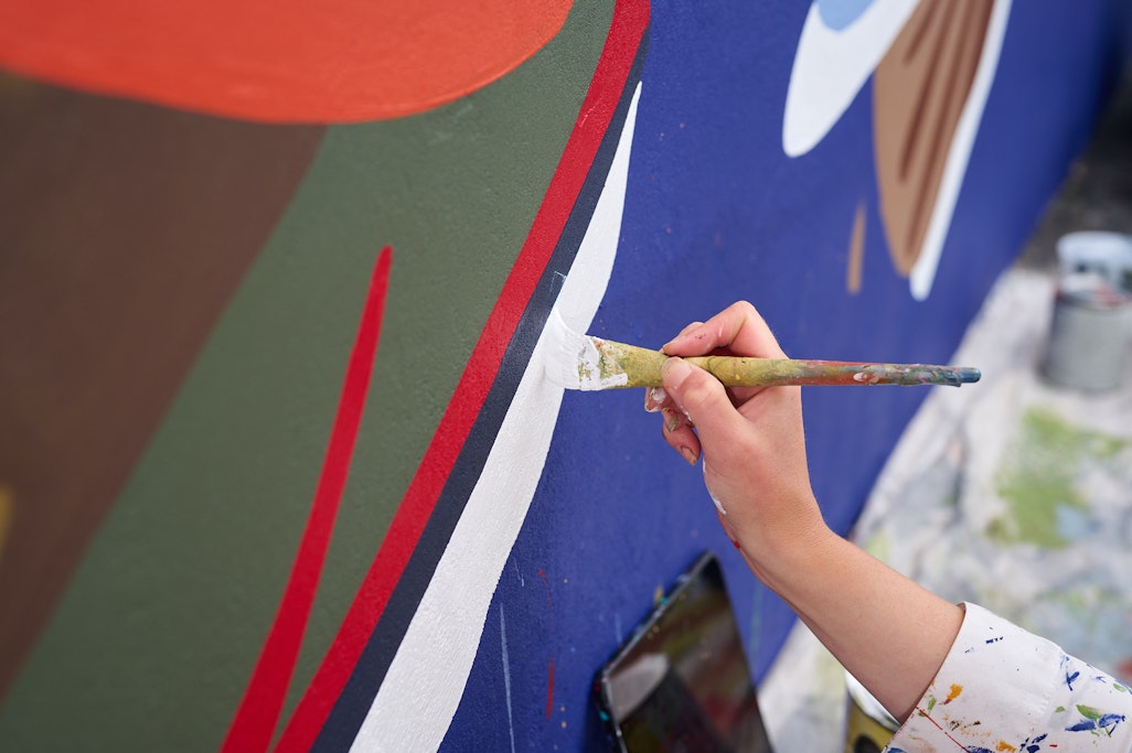 a painter’s hand applying white paint to a mural with splashes of red, blue and orange in the background
