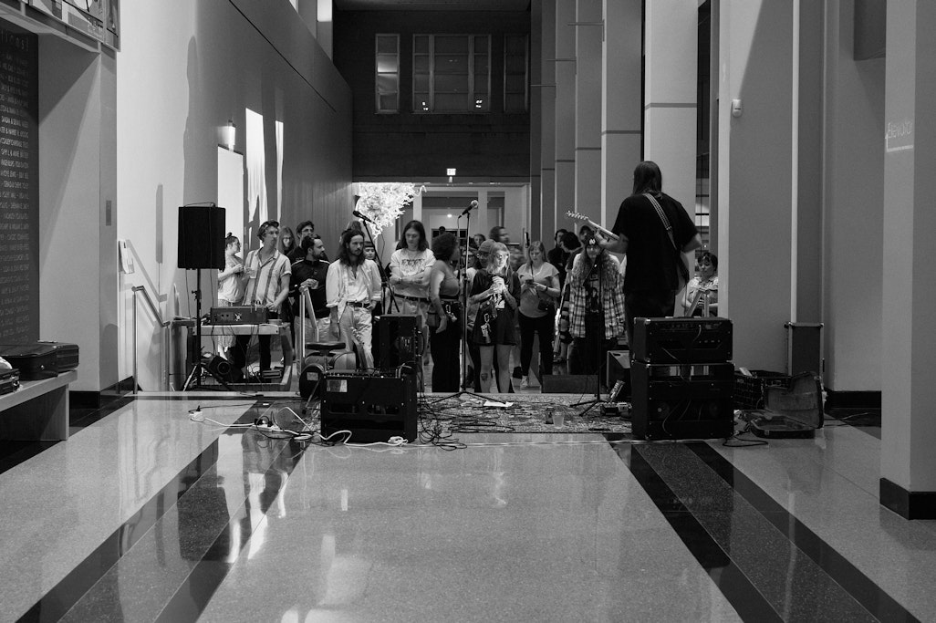 wide shot down a long hall from far behind the stage where a musician is playing, the crowd at a Gray Duck rock music show, at Fine Arts Center